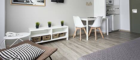 A modern, well-lit room with a dining area, a TV mounted on the wall, and a kitchenette.