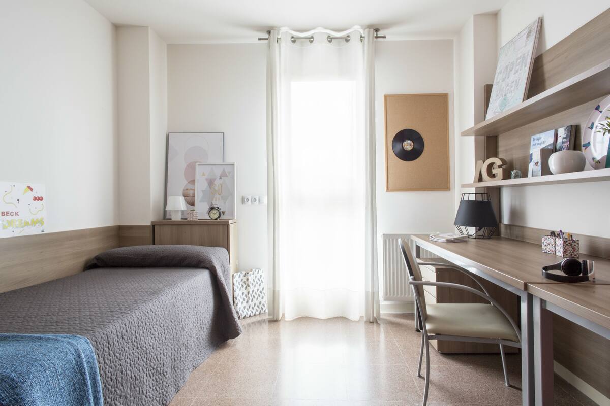 A bedroom with a bed, a desk, and a chair. There is a window with curtains, a radiator, and wall decorations.