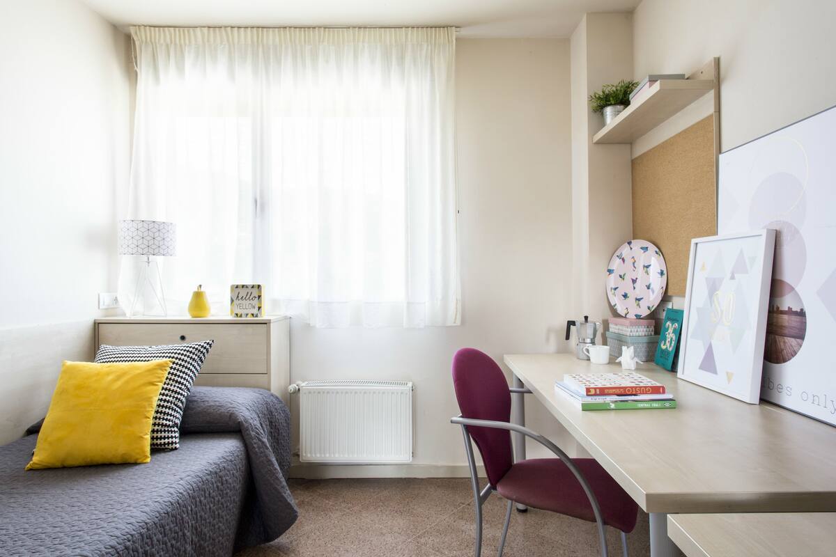 A bedroom with a bed, a desk, a chair, a radiator, a window with curtains, and some wall decorations.