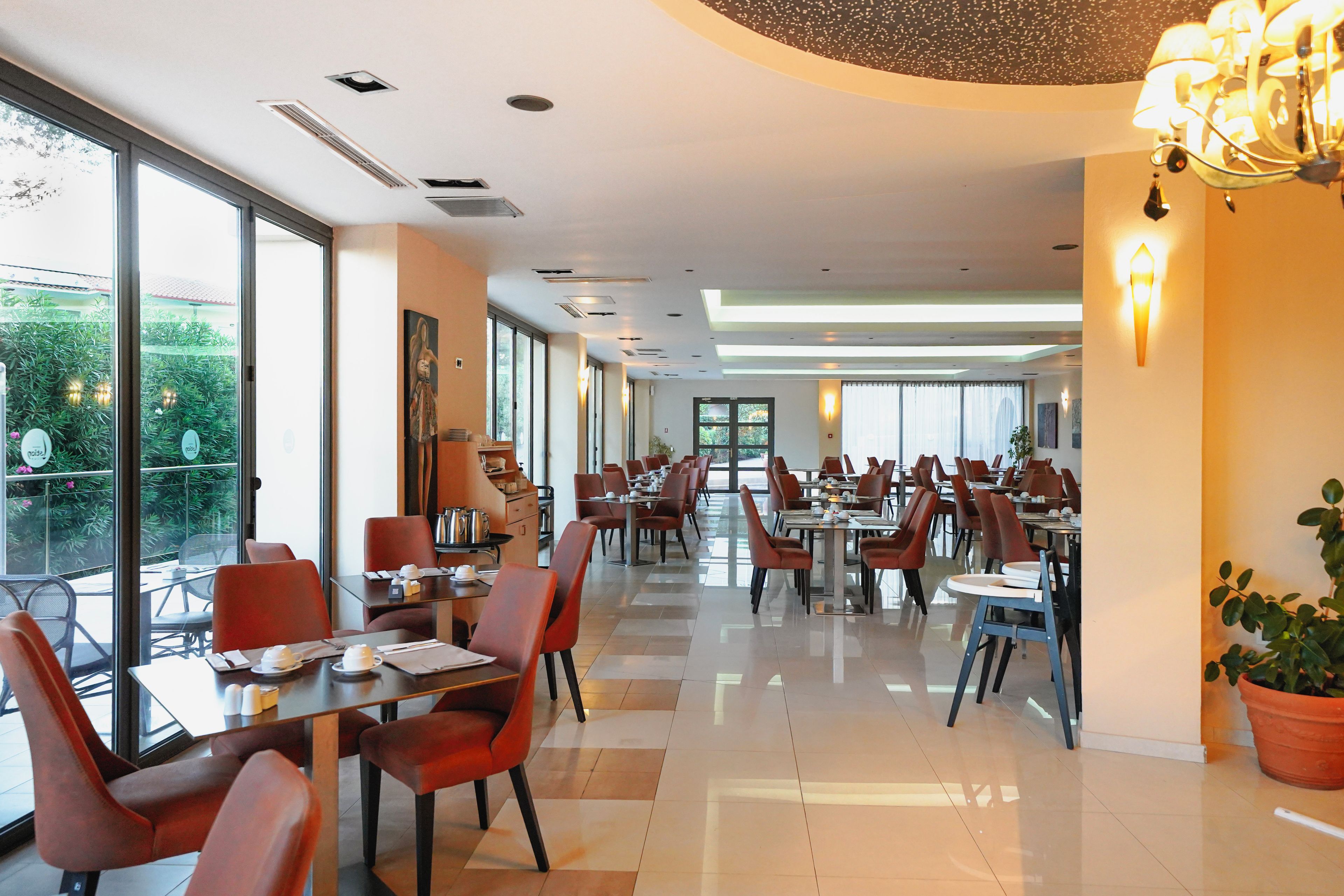 Un comedor de hotel con mesas y sillas, ventanas amplias y vistas al exterior con vegetación.