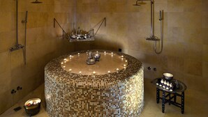 Eine runde Badewanne mit Mosaikfliesen, ein kleines Tischchen und Kerzen, umgeben von Duscharmaturen.