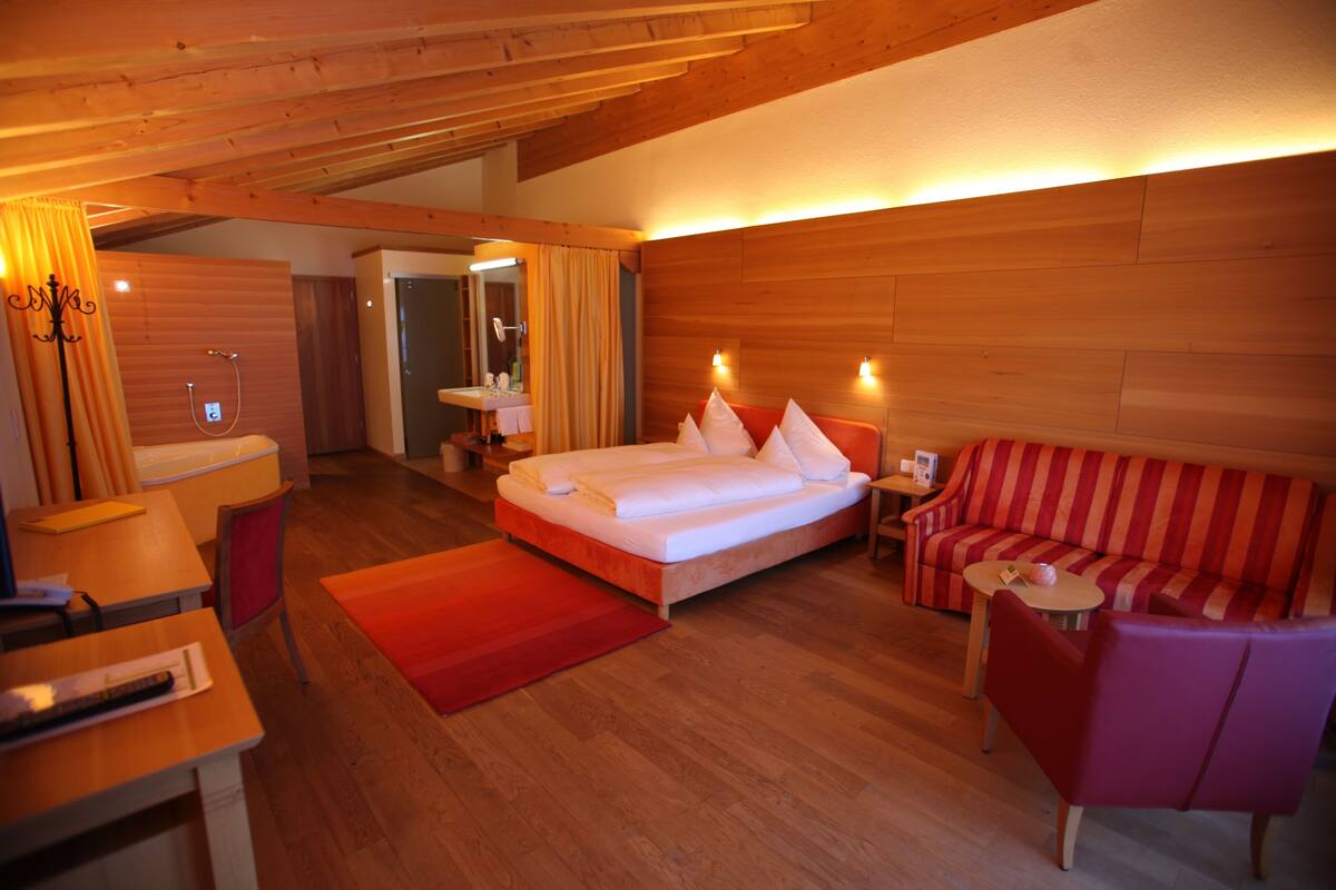 A hotel room with a wooden ceiling, a bed with white linens, a red-striped sofa, a small table, and a desk with a chair.