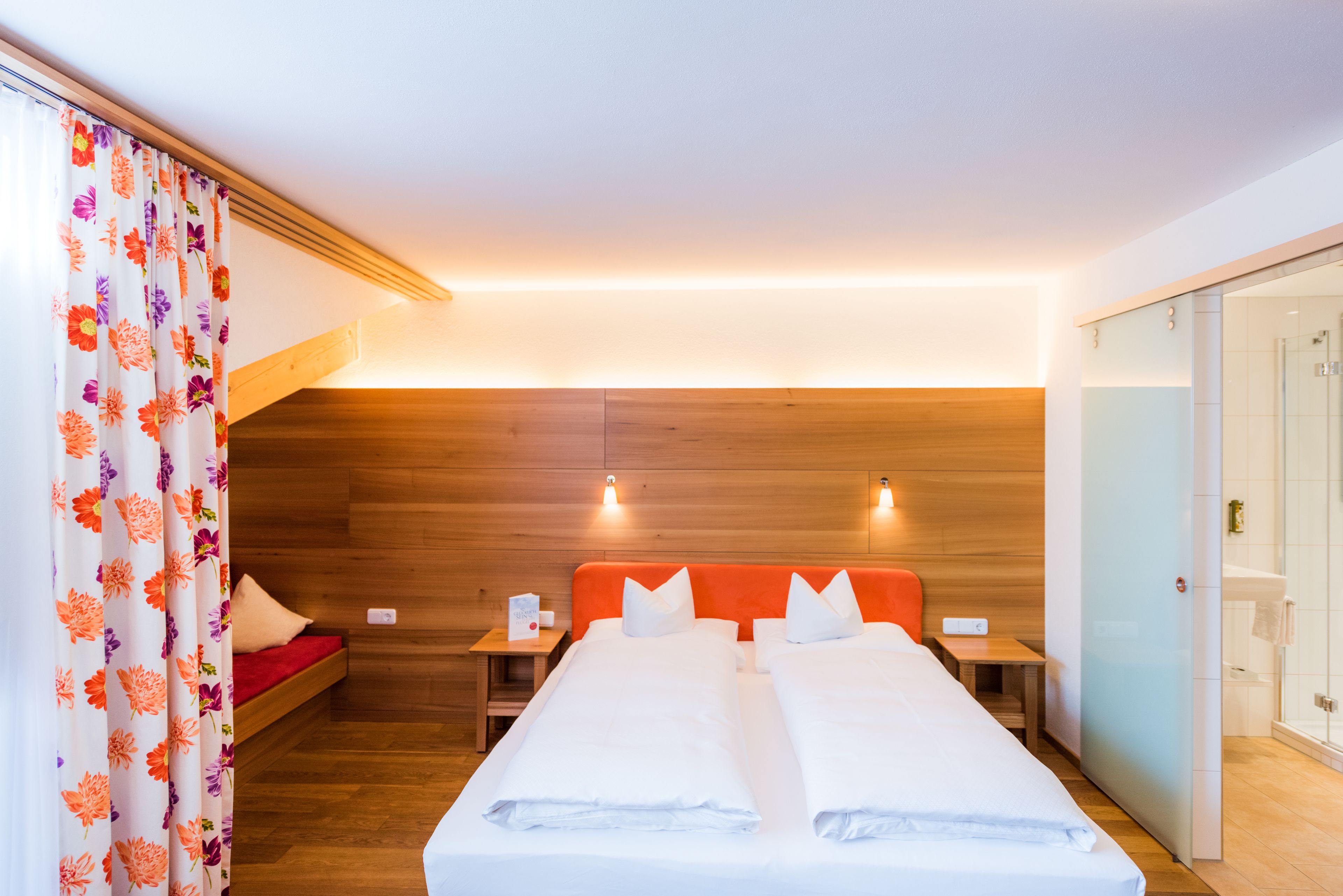 A hotel room with a wooden ceiling, a bed with white linens, a red headboard, and a small bedside table with a lamp.