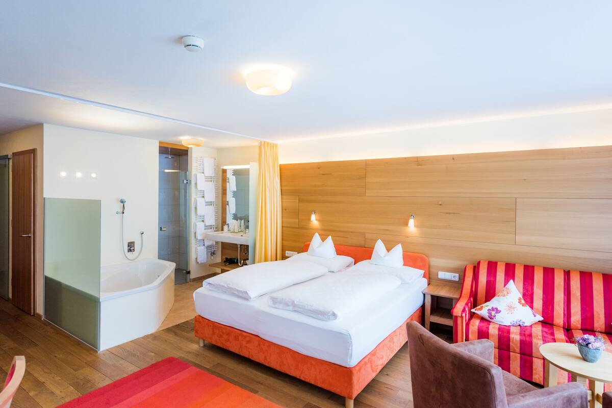 A modern hotel room with a large bed, a separate bathroom, a wooden headboard, and a small seating area with a striped sofa.