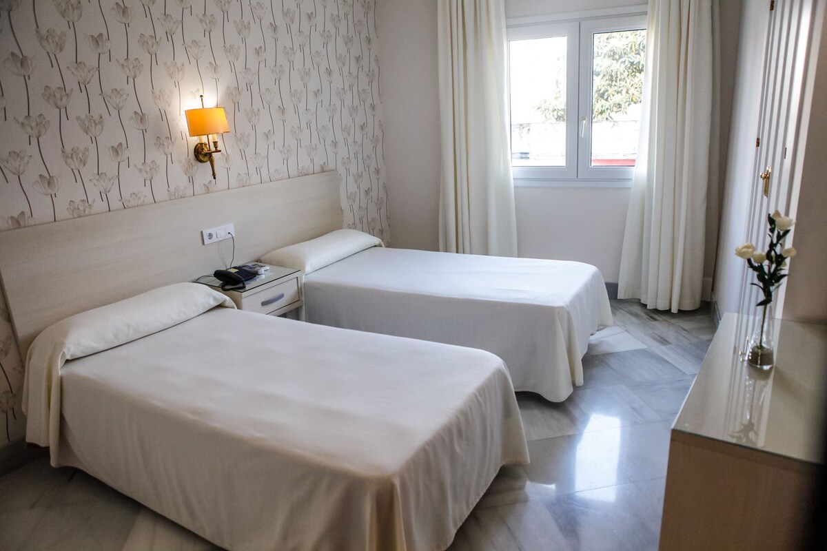 Une chambre d’hôtel avec deux lits, une table de chevet, une lampe, une fenêtre avec des rideaux et un vase avec des fleurs.