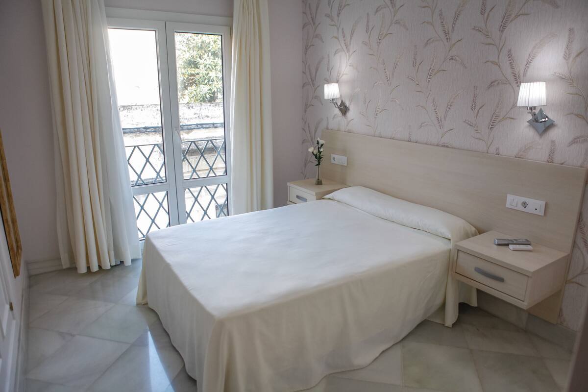 Une chambre d’hôtel avec un grand lit, deux tables de chevet, une petite table, une lampe et une vue à travers une porte vitrée.