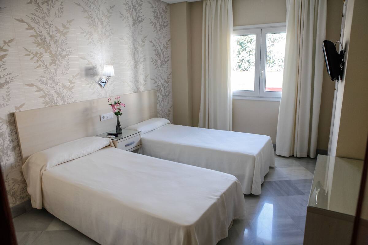 Une chambre d’hôtel avec deux lits, une table de chevet avec une lampe, une fenêtre avec des rideaux et un téléviseur fixé au mur.