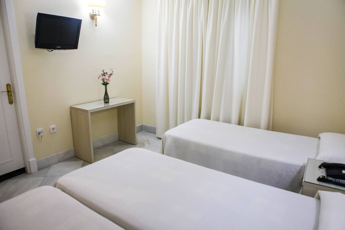 Une chambre d’hôtel avec deux lits, un téléviseur fixé au mur, un petit bureau agrémenté d’un vase de fleurs et un rideau.