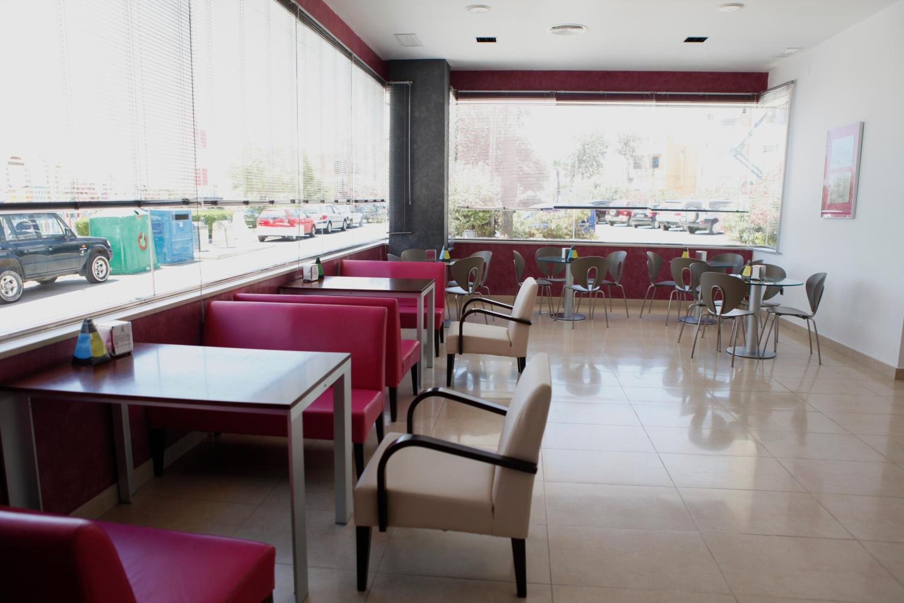 A restaurant with red booths and tables, large windows with a view of parked cars and buildings outside.