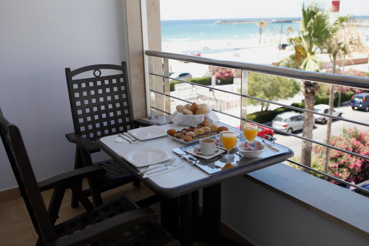 Mesa de balcón con alimentos para el desayuno, una silla y vista a la playa y al océano.