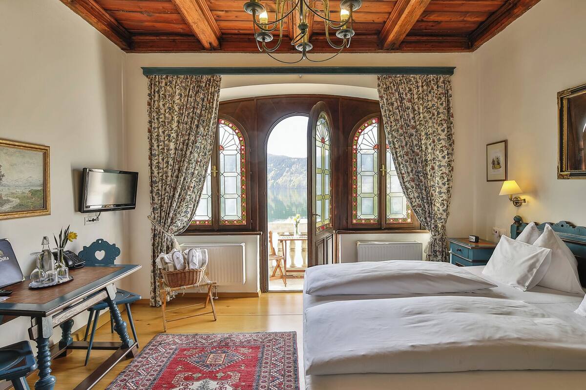 Une chambre à coucher avec un grand lit, un bureau avec un ordinateur, une chaise et une vue sur un lac, visible à travers des fenêtres en arc de cercle.