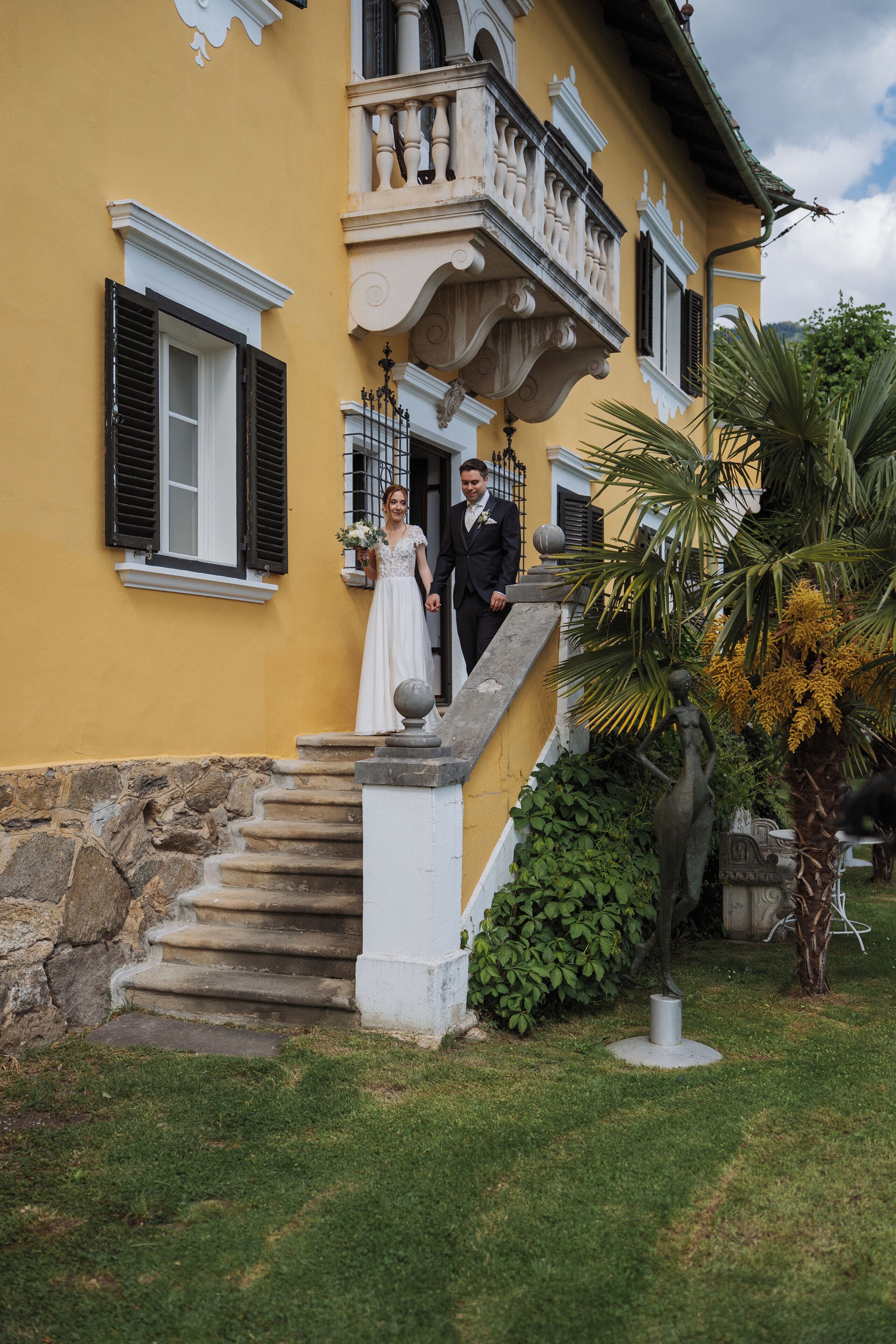 A couple on a stone staircase outside a yellow building with a balcony.