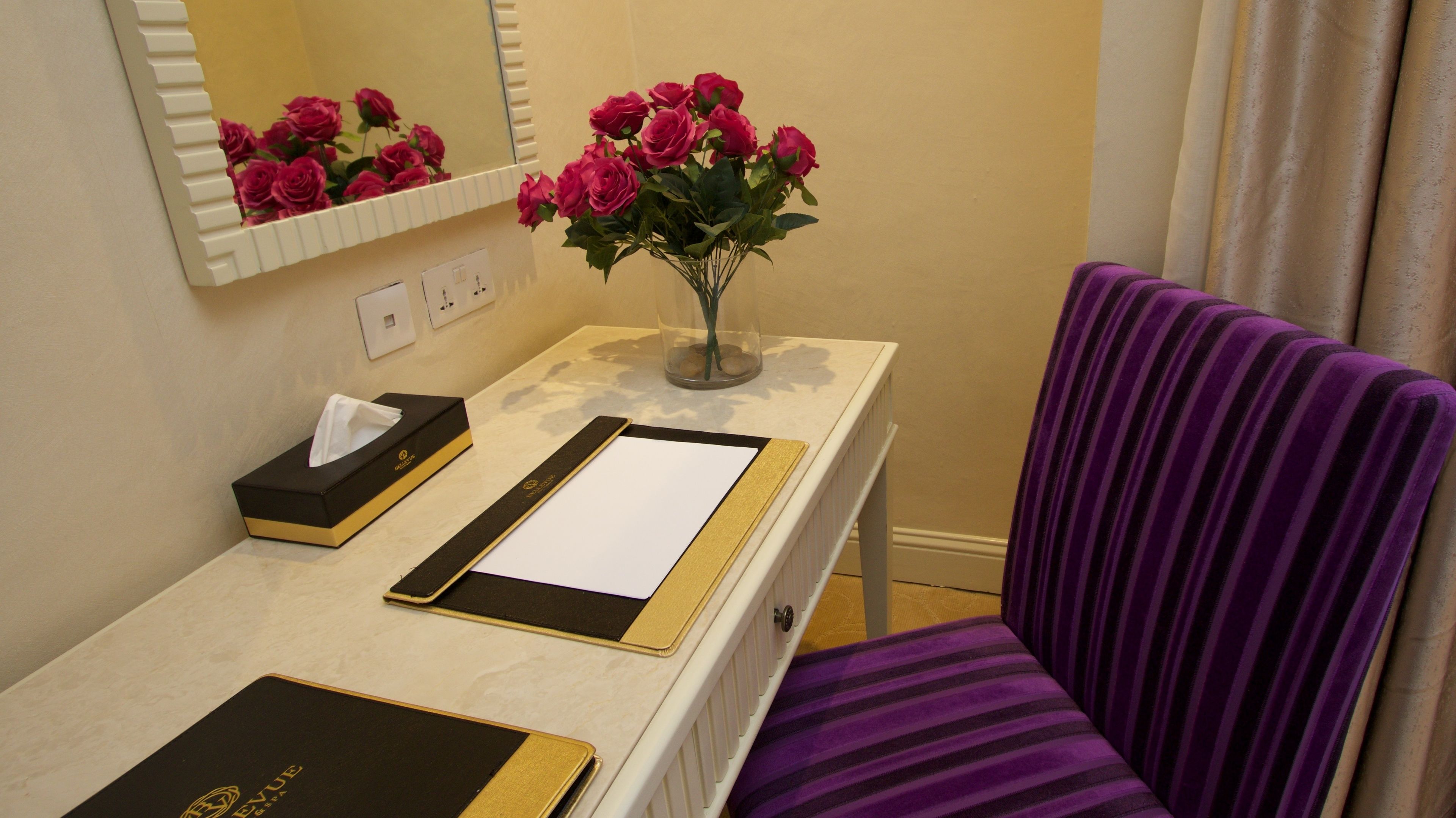 A hotel room with a desk, a vase of pink flowers, a tissue box, a black notebook, and a purple striped chair.
