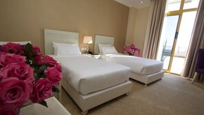 A hotel room with two beds, a nightstand with a lamp, a vase of pink flowers, and a view of the outside through a large window. - Bellevue Hotel and Spa (Addis Ababa)