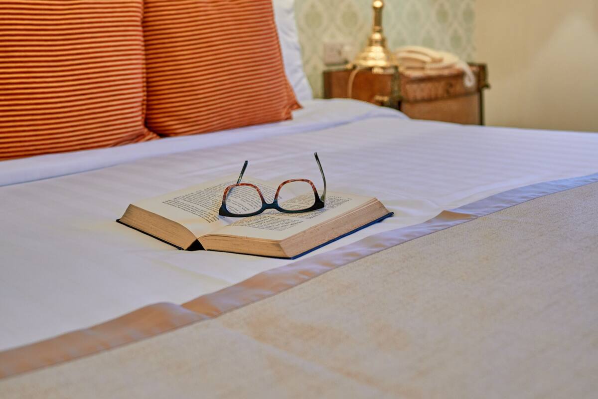 Una cama bien tendida con almohadas naranjas, una lámpara de noche y un libro abierto con gafas encima.