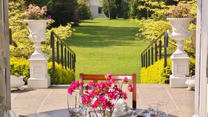 Mesa de comedor preparada para seis personas, con flores rosadas, vasos y cubiertos, con vistas a un césped y jardín cuidados.