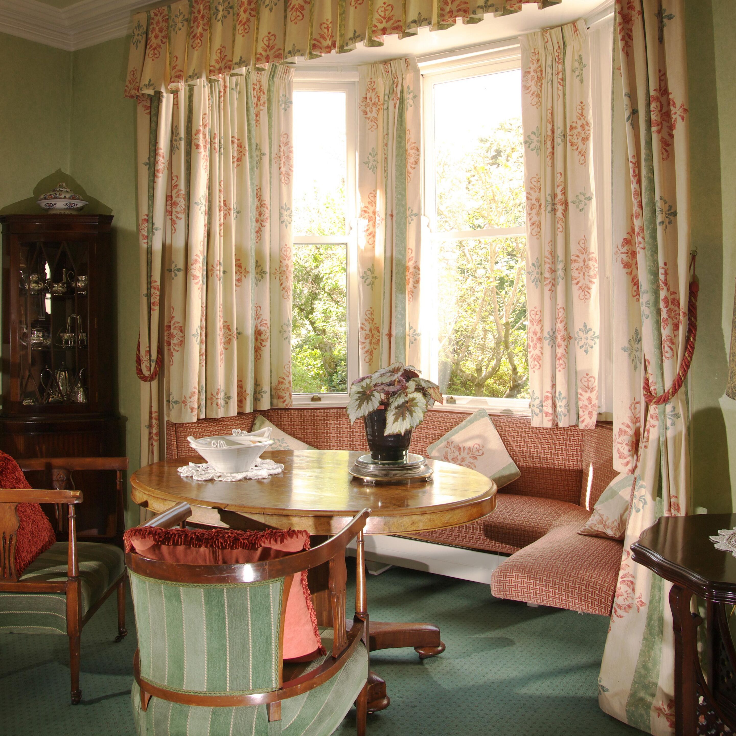 A cozy room with a round wooden table, a bench with cushions, a cabinet with glass doors, and a window with floral curtains.