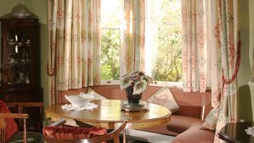 A cozy room with a round wooden table, a bench with cushions, a cabinet with glass doors, and a window with floral curtains.