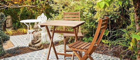 A wooden table and chair set on a patterned tile floor, surrounded by lush greenery and a statue.