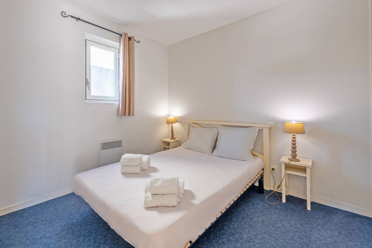 A neatly made bed with white linens and towels, two bedside tables with lamps, a window with curtains, and a radiator.