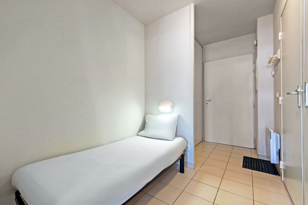 A small, single-bed room with a bedside table, a wall-mounted light, and a door leading to another room.