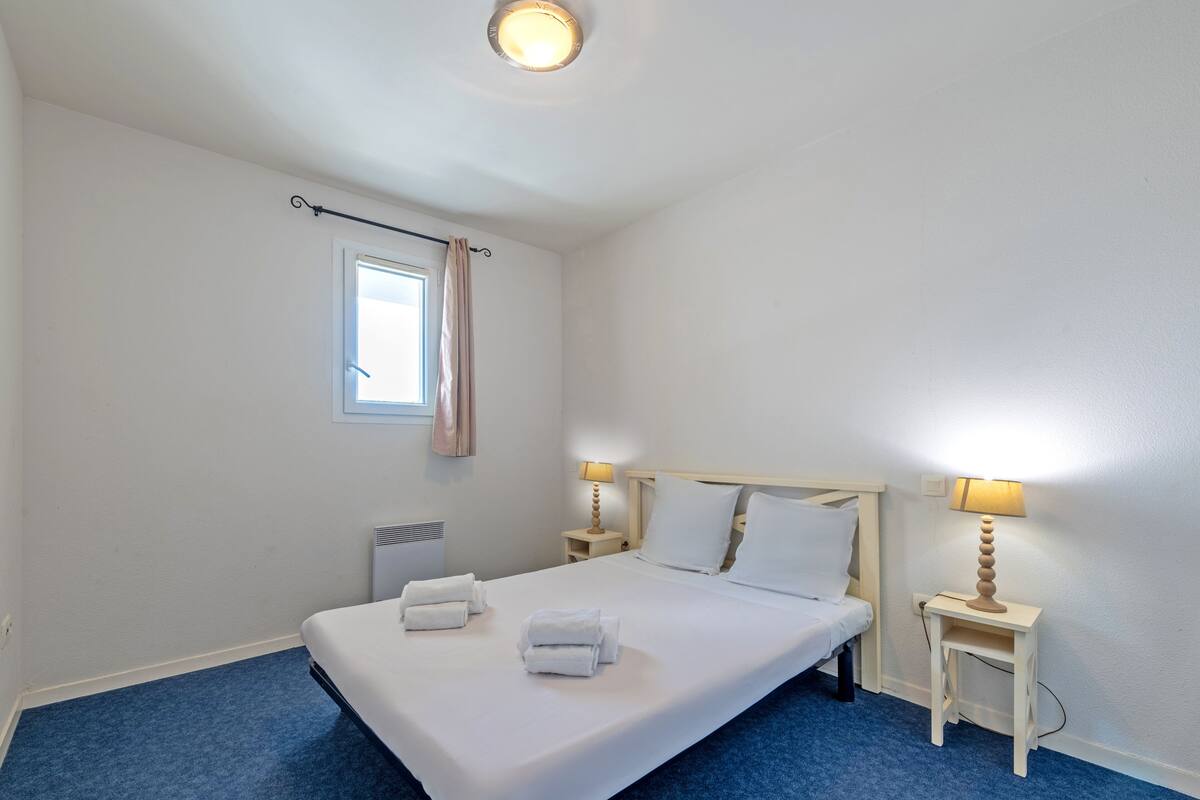 A neatly made bed with white linens and towels, a window with a curtain, bedside lamps, and a radiator.