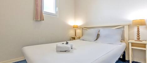 Una cama bien hecha con sábanas blancas, dos lámparas de noche y una ventana con cortina.