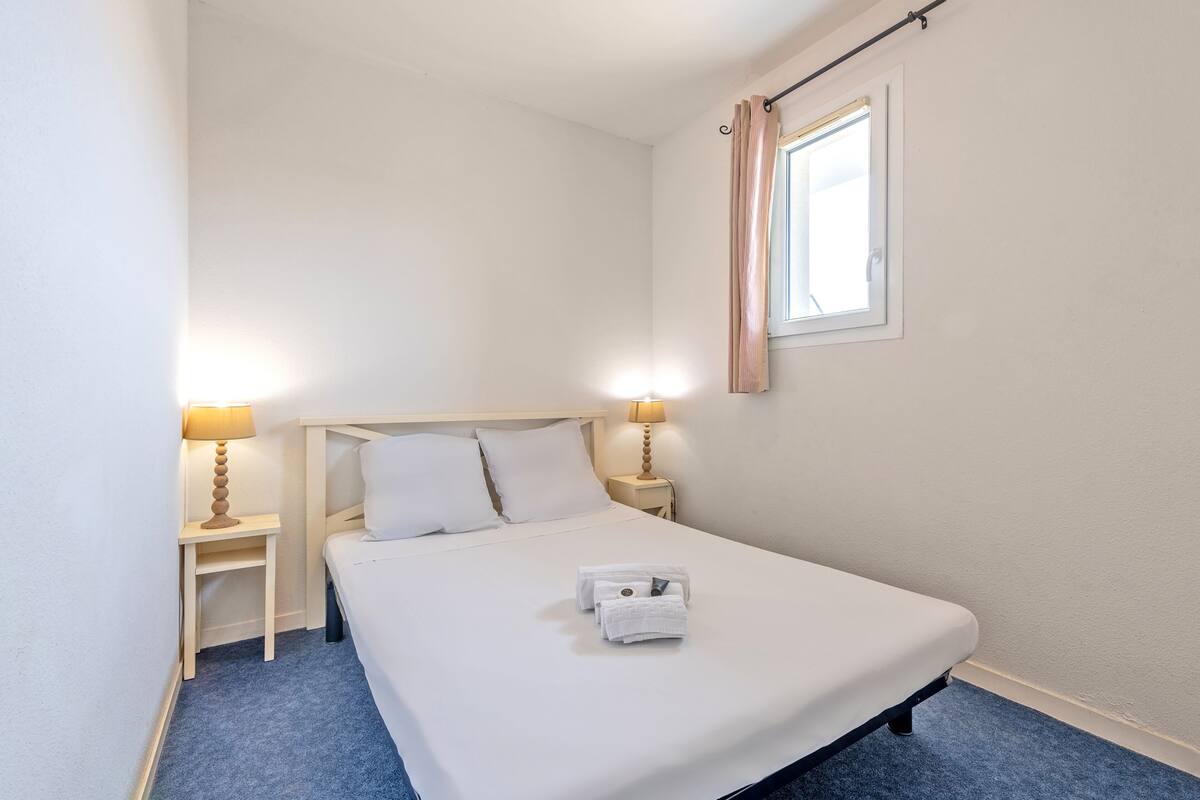 A single-bed room with a wooden headboard, two bedside lamps, a window with curtains, and a blue carpet.