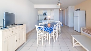 A modern kitchen with white cabinets, a dining table with chairs, a TV on a stand, and a refrigerator. - Vacancéole Le Domaine de Bertheaume (Plougonvelin)