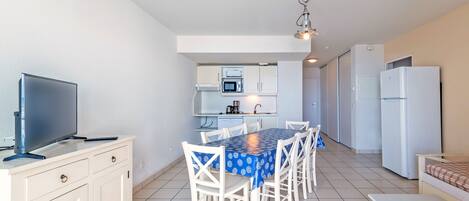 A modern kitchen with white cabinets, a dining table with chairs, a TV on a stand, and a refrigerator.