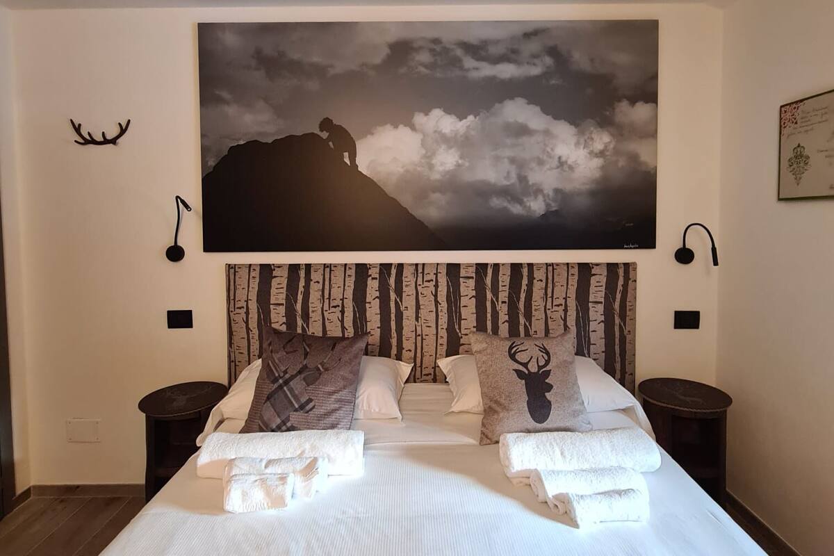 Une chambre à coucher avec un grand lit, une tête de lit ornée d’un motif de forêt, un tableau représentant une montagne et deux tables de chevet avec des lampes.