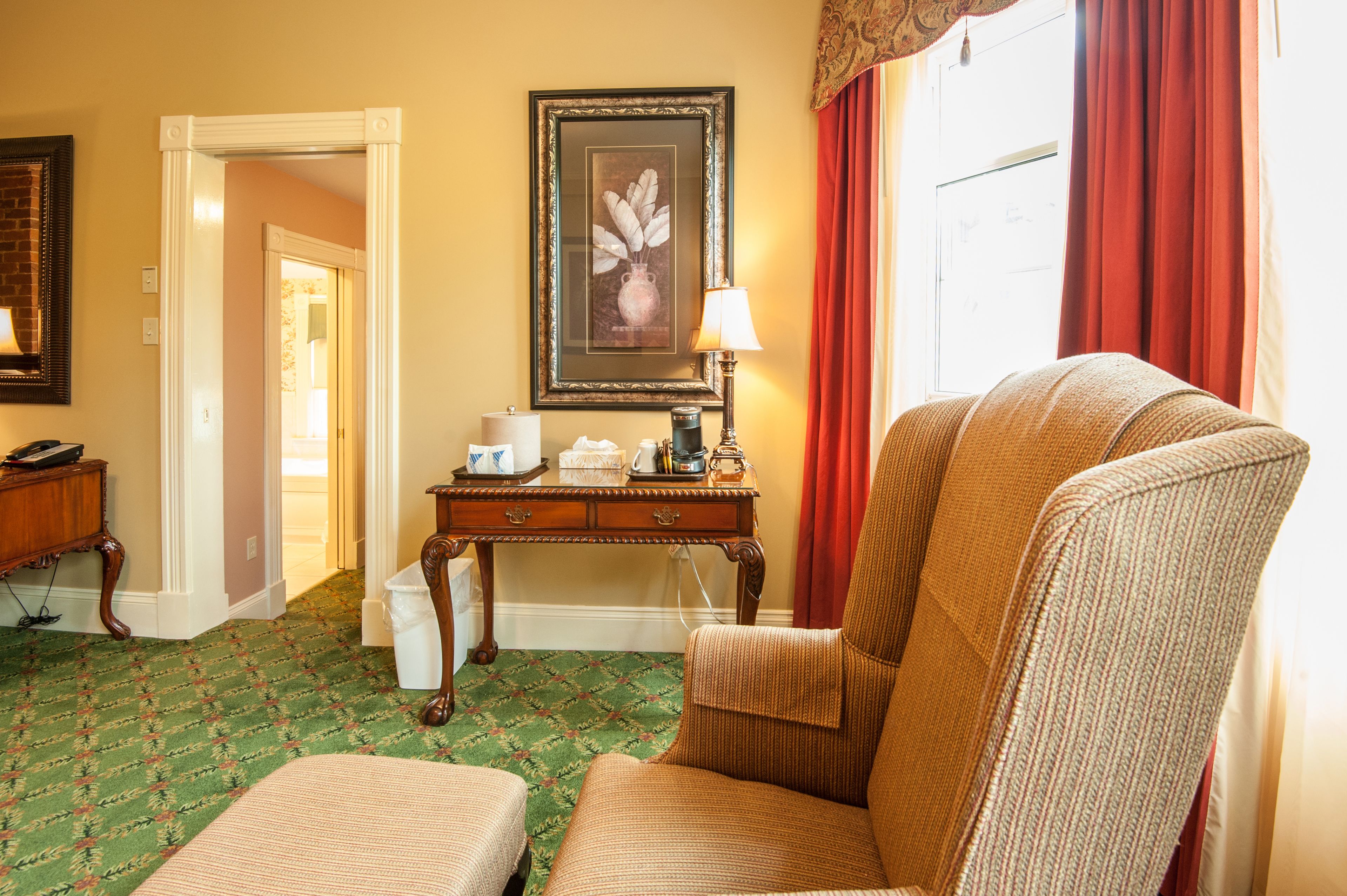 A hotel room with a green patterned carpet, a wooden desk with a lamp, a framed picture on the wall, and a window with red curtains.