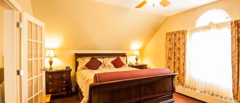 A bedroom with a large bed, two bedside tables with lamps, a ceiling fan, and a window with curtains.