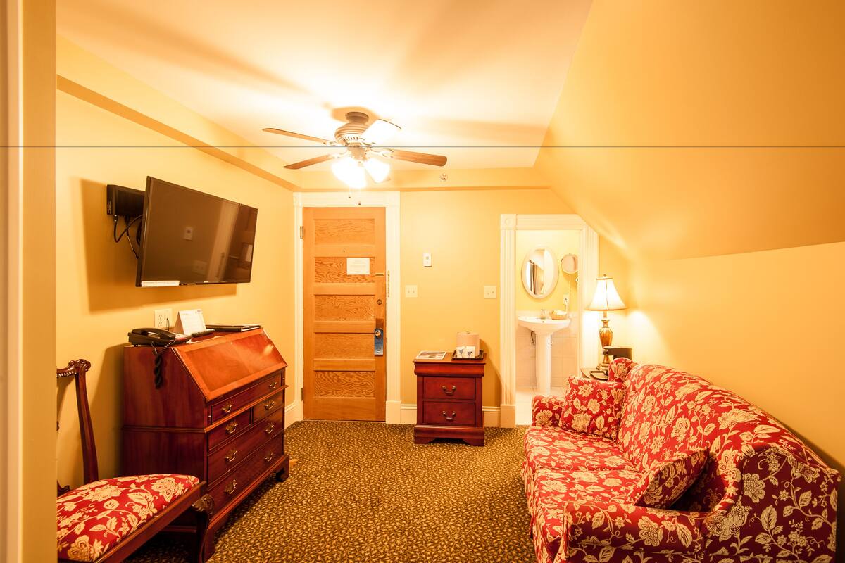 Ein Hotelzimmer mit einem hölzernen Schminktisch, einem roten Blumsofa, einem Deckenventilator, einem an der Wand montierten Fernseher und einer Tür zum Badezimmer.