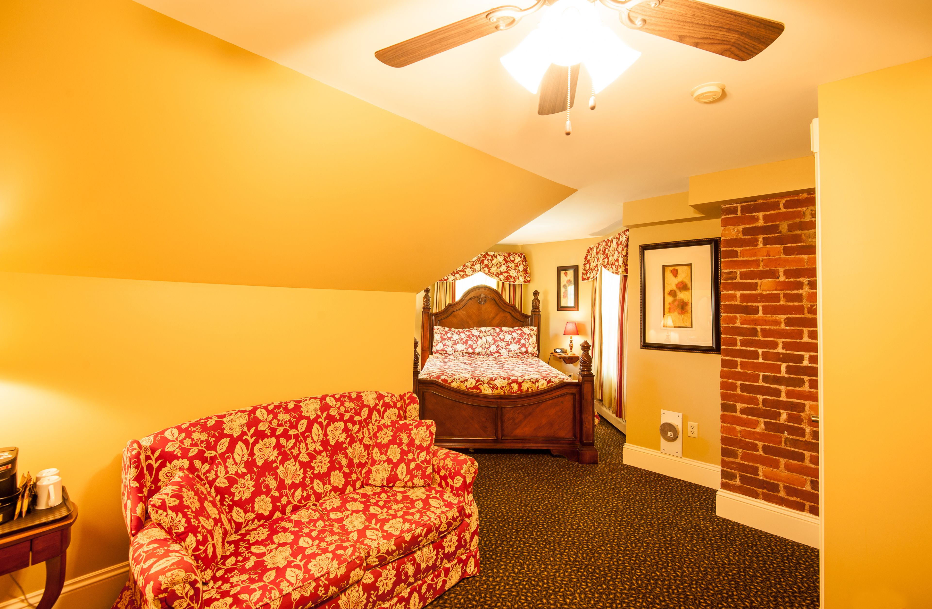 A bedroom with a bed, a red floral sofa, a ceiling fan, a brick wall, and a small table with a lamp.