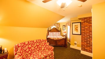 A bedroom with a bed, a red floral sofa, a ceiling fan, a brick wall, and a small table with a lamp.