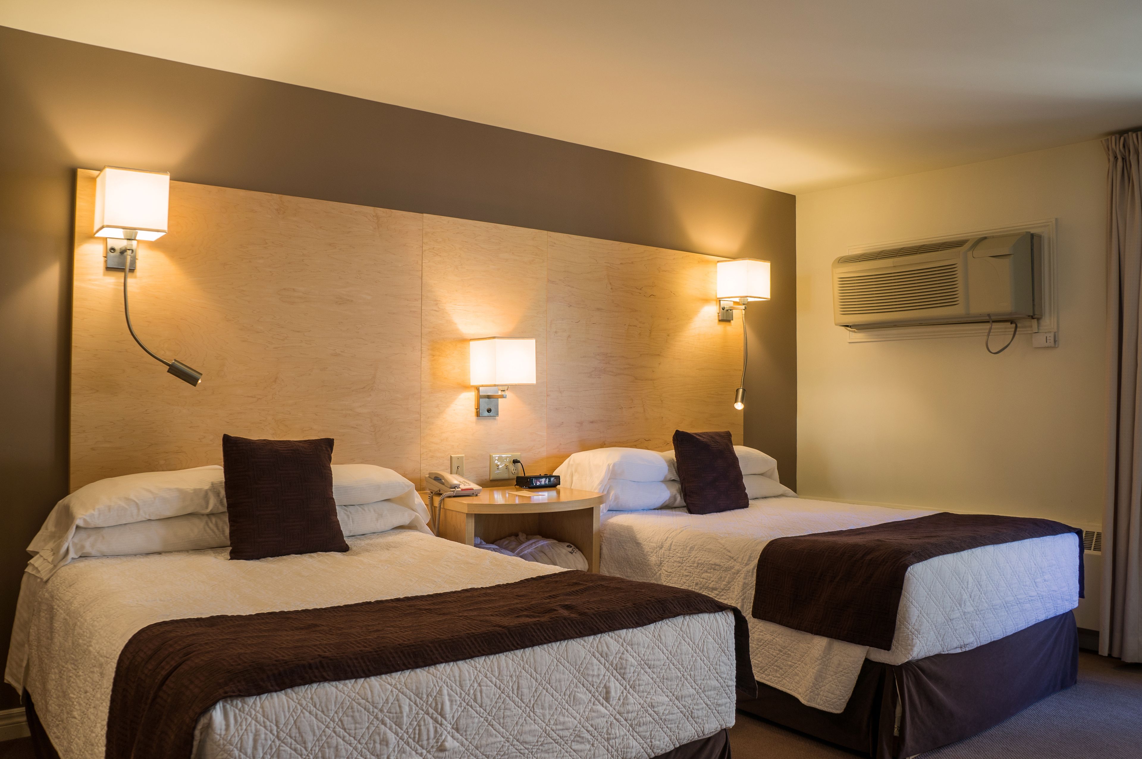 Habitación de hotel con dos camas, cada una con almohada y manta. Se observa un mesita de noche con lámpara y teléfono. Una unidad de aire acondicionado está montada en la pared.