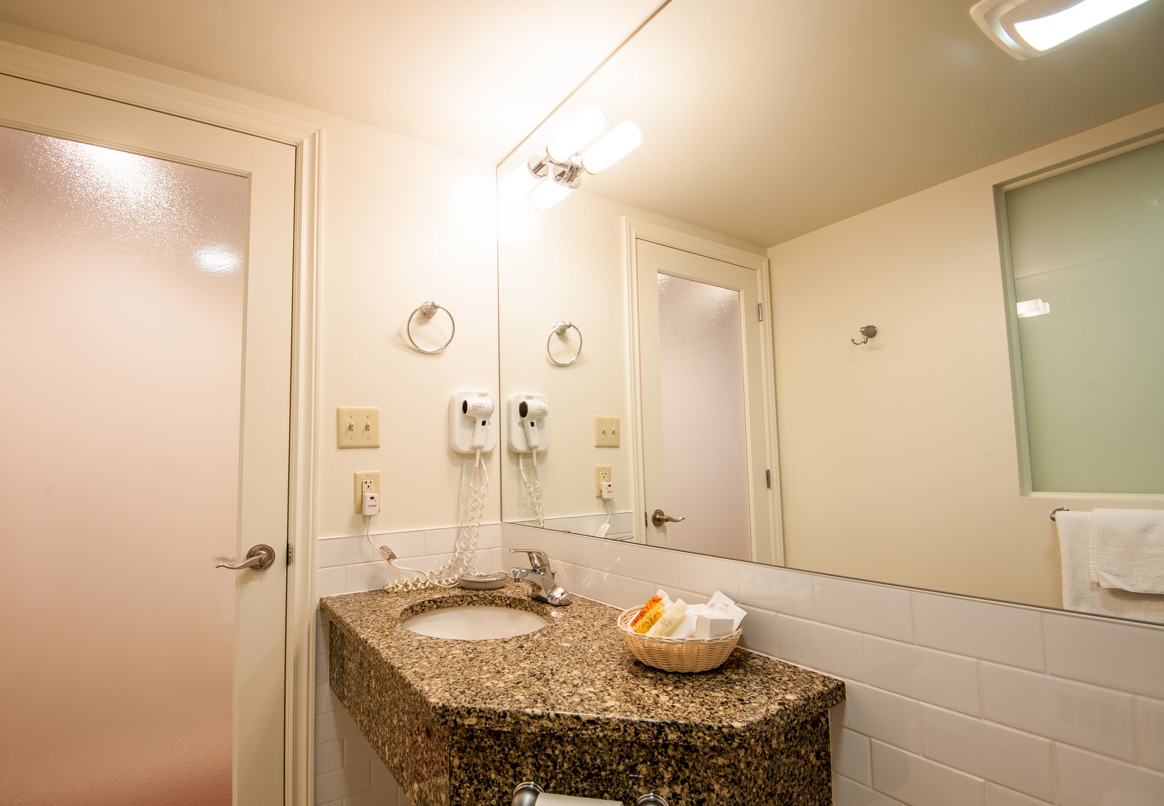A bathroom with a granite countertop sink, a mirror, a towel rack, and a basket of toiletries.