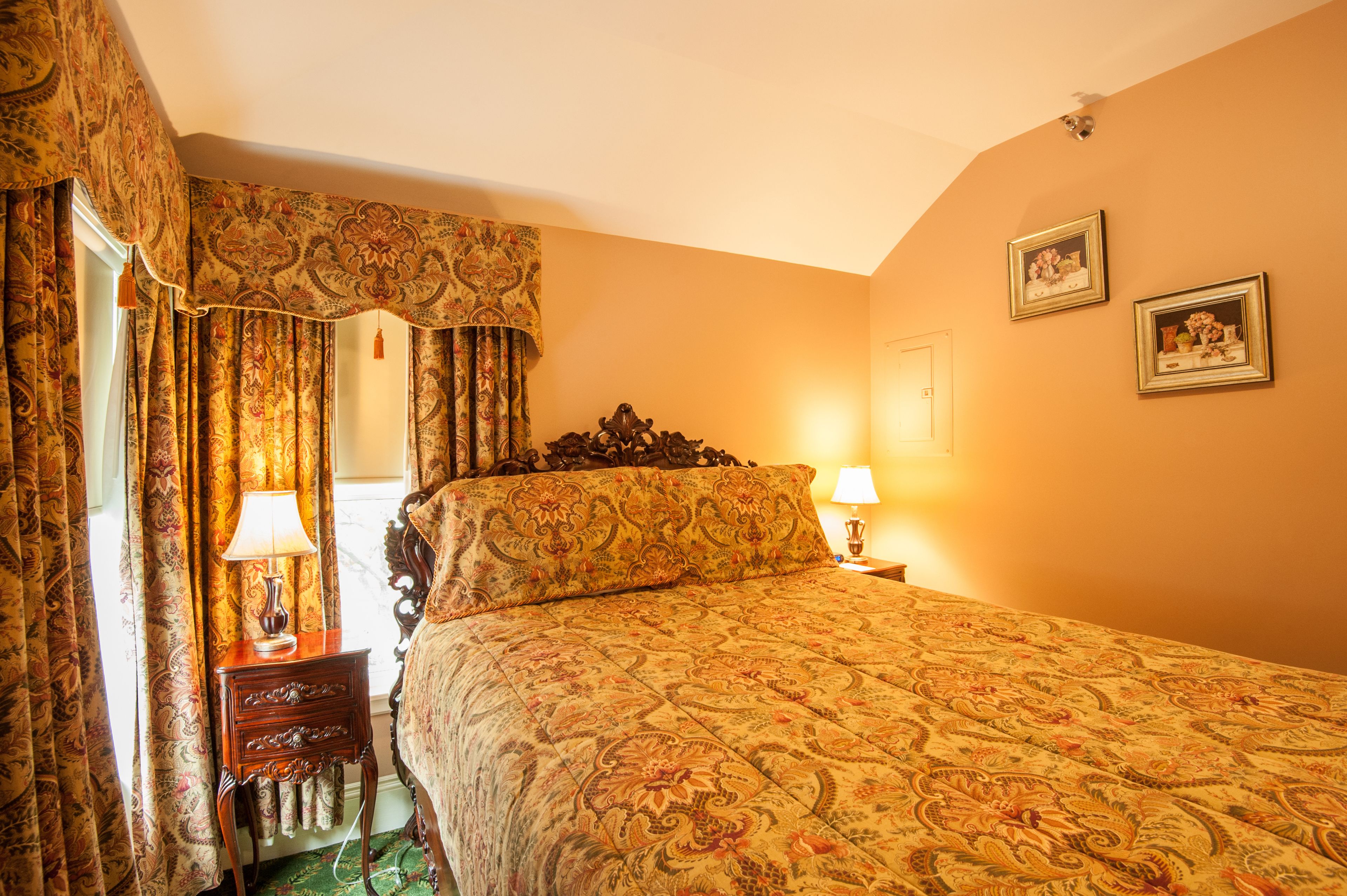Een hotelkamer met een groot bed, sierlijke gordijnen, een nachtkastje met een lamp en twee ingelijste foto's aan de muur.
