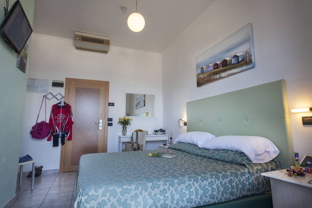 Une chambre d’hôtel avec un lit, un bureau et une chaise, un miroir, un tableau accroché au mur et une porte.