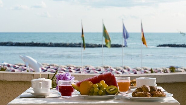 A breakfast table with fruit, pastries, and drinks, overlooking a beach with flags. - Hotel San Giorgio Savoia (Bellaria-Igea Marina)