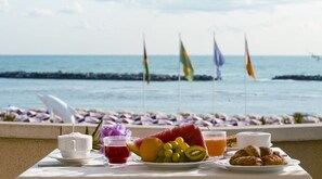 A breakfast table with fruit, pastries, and drinks, overlooking a beach with flags. - Hotel San Giorgio Savoia (Bellaria-Igea Marina)