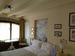 Une chambre à coucher avec un lit, une chaise, une petite table et une lampe. Il y a des rideaux à la fenêtre et un tableau au mur.