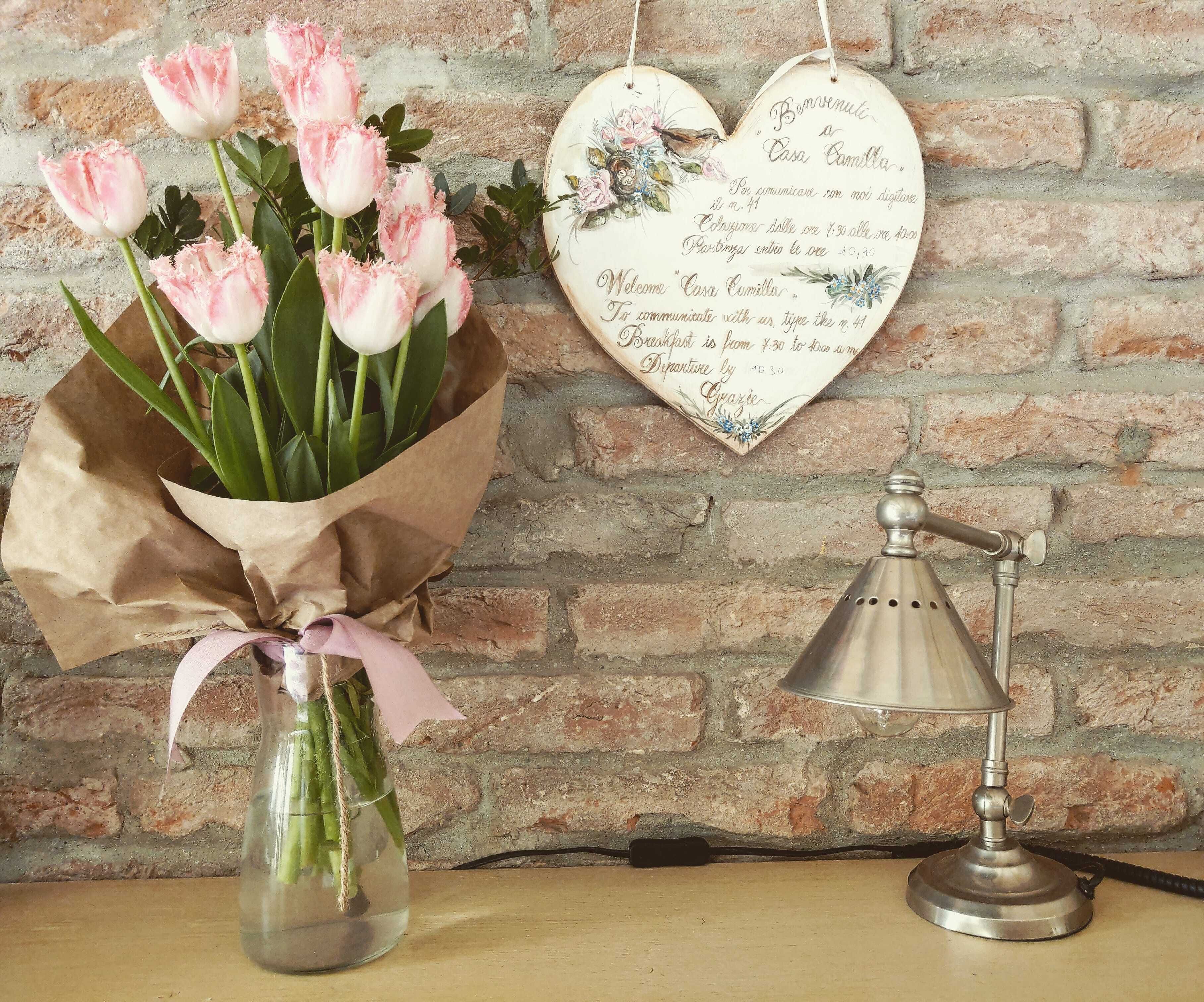 Un ramo de tulipanes rosados en un florero de cristal con envoltorio de papel marrón y una cinta rosa, una lámpara de pared de estilo vintage, y un letrero decorativo con forma de corazón con un mensaje.