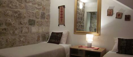 A room with two single beds, a stone wall, a mirror, and a small table with a lamp.
