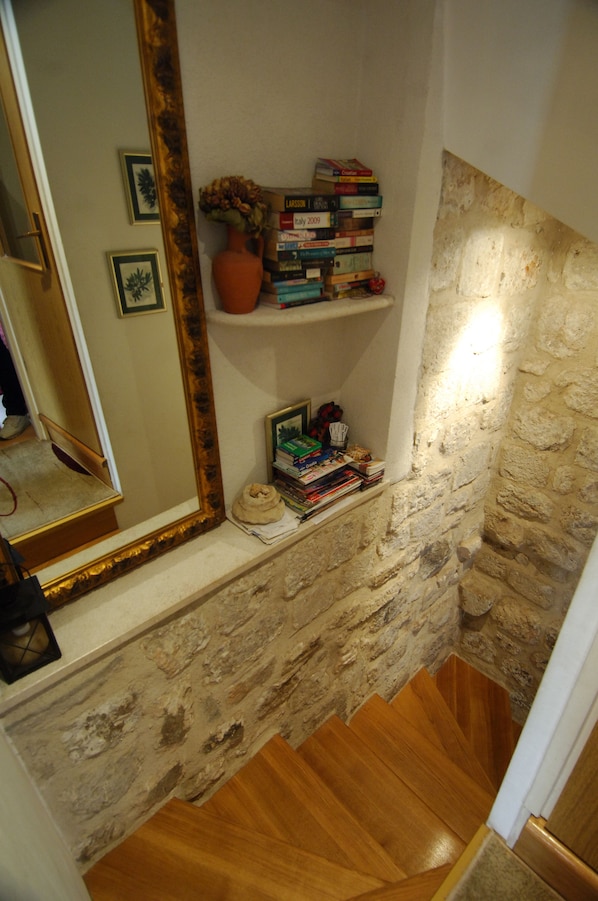 A staircase with wooden steps and a stone wall. There is a mirror on the left and a shelf with books and a vase on the right.