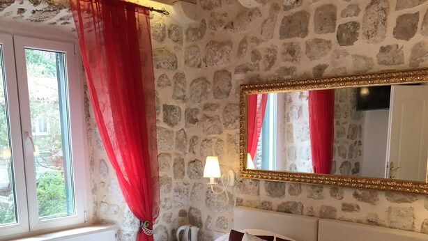 Un dormitorio con pared de piedra, una cama grande con ropa de cama blanca y roja, una mesita de noche con una lámpara y una ventana con cortinas rojas.