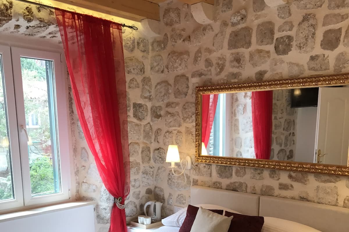 A bedroom with a stone wall, a large bed with white and red bedding, a bedside table with a lamp, and a window with red curtains.