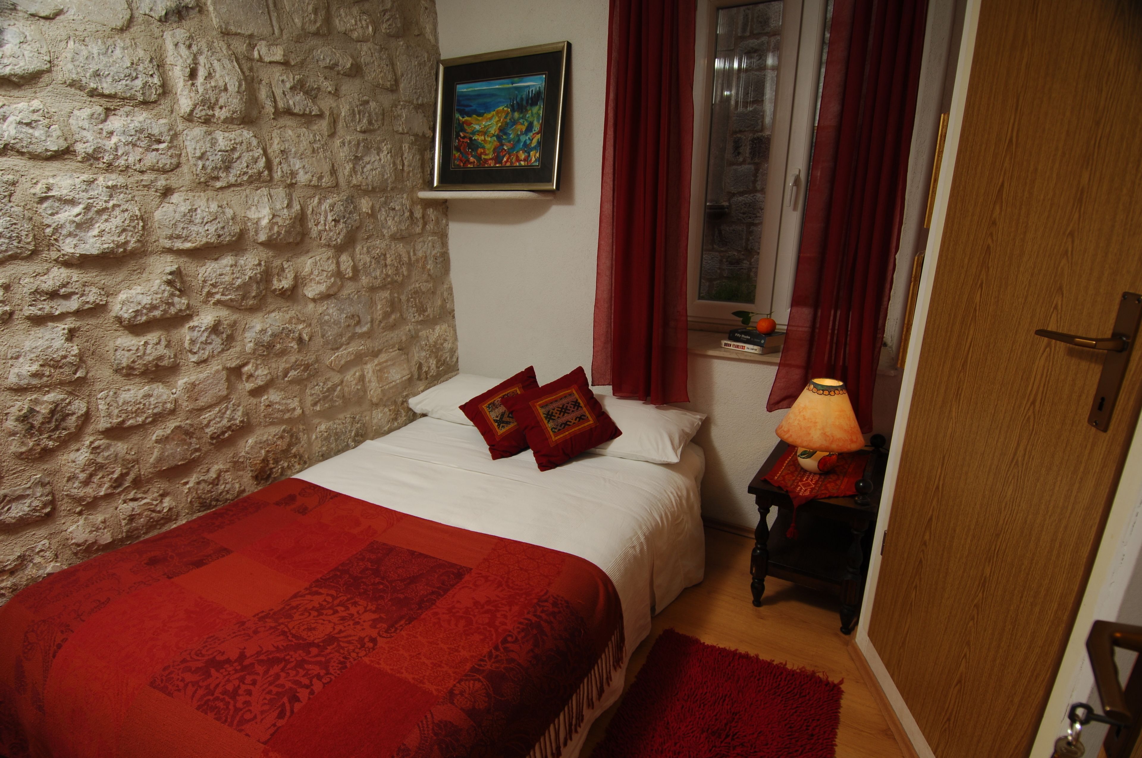 Une chambre avec un mur en pierre, un lit avec une literie rouge, un tableau accroché au mur et une fenêtre ornée de rideaux rouges.