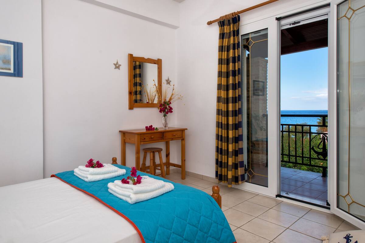 Un dormitorio con cama, escritorio, espejo y vista al mar a través de una puerta corrediza de vidrio.